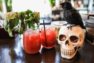 2 cocktails on a table next to a fake skull with a black crow sitting on top